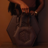 A person holds two dark brown, hexagon-shaped bags of different sizes, one with a prominent logo. The person wears a ring and bracelet and stands against a warm, brown background.