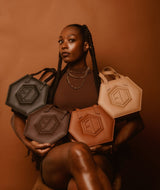 A woman with long braids is seated against a brown backdrop, holding four hexagonal handbags in various shades of brown. Each bag features a circular embossed logo. She wears a brown top and layered necklaces, gazing confidently at the camera.