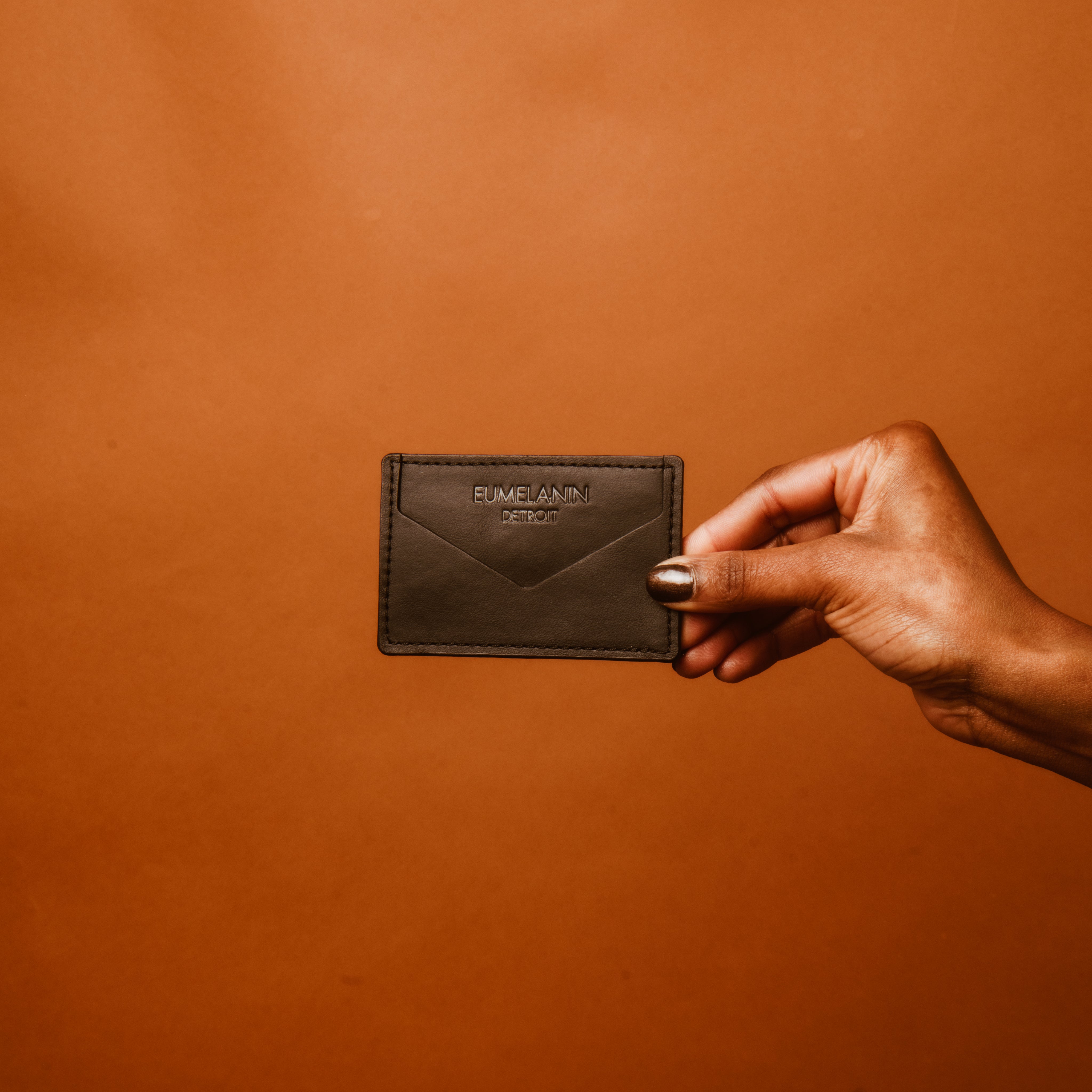 A hand, nails painted dark with the richness of eumelanin, holds a dark brown leather wallet against a solid brown backdrop. The wallet is embossed with text—a perfect accessory to shop in style.