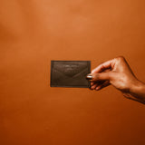 A hand, nails painted dark with the richness of eumelanin, holds a dark brown leather wallet against a solid brown backdrop. The wallet is embossed with text—a perfect accessory to shop in style.