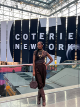 A person in stylish dark clothing, rich in eumelanin hues, and boots stands on a glass walkway, holding a brown handbag. In front of large banners that read Coterie Network, the scene is an indoor venue with pink carpeted areas below—a perfect backdrop for a chic shopping experience.