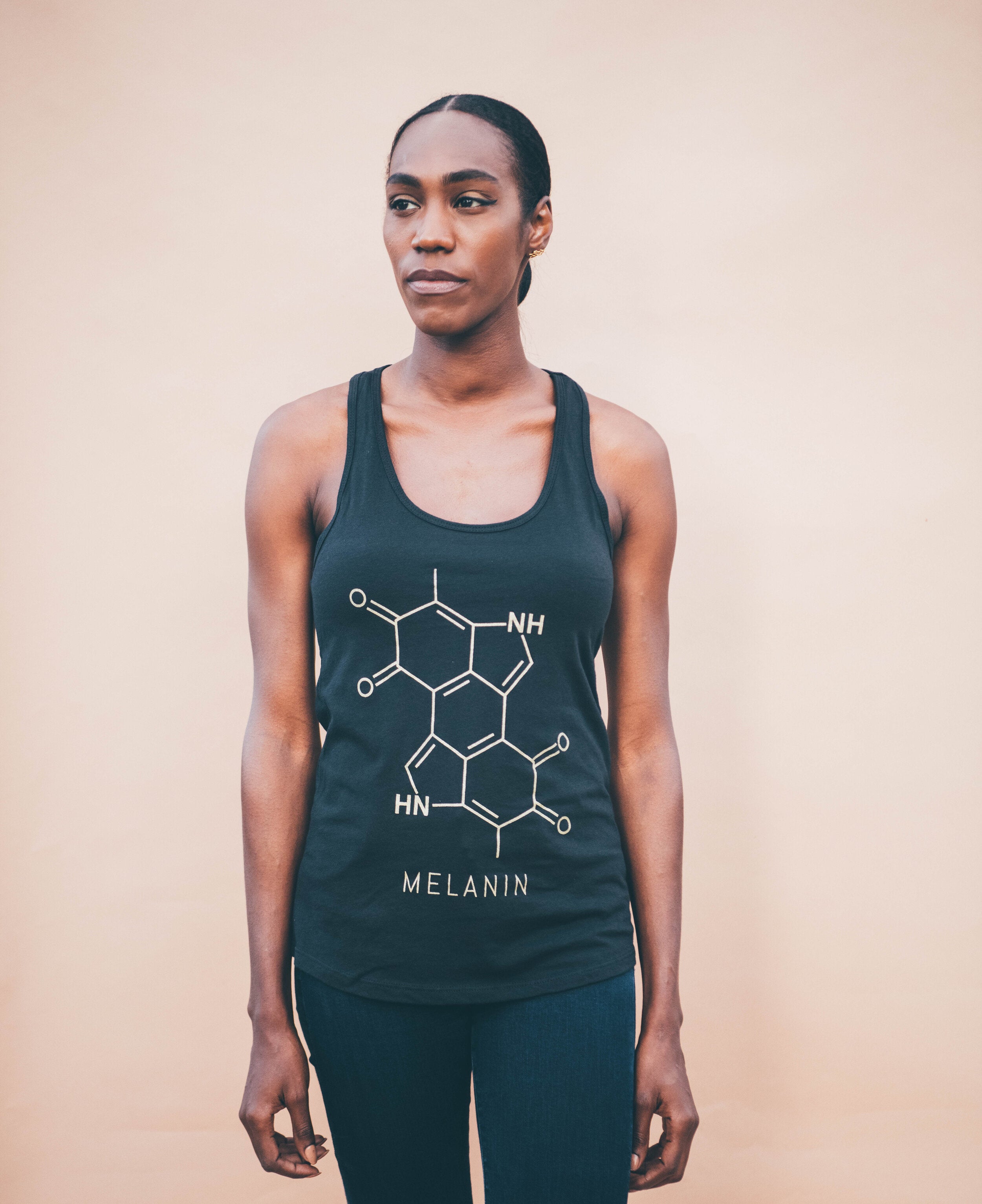 A woman poses against a plain background, sporting a black sleeveless top with a gold print of the melanin molecule and MELANIN below it. Her long hair is tied back, and she completes the look with jeans.