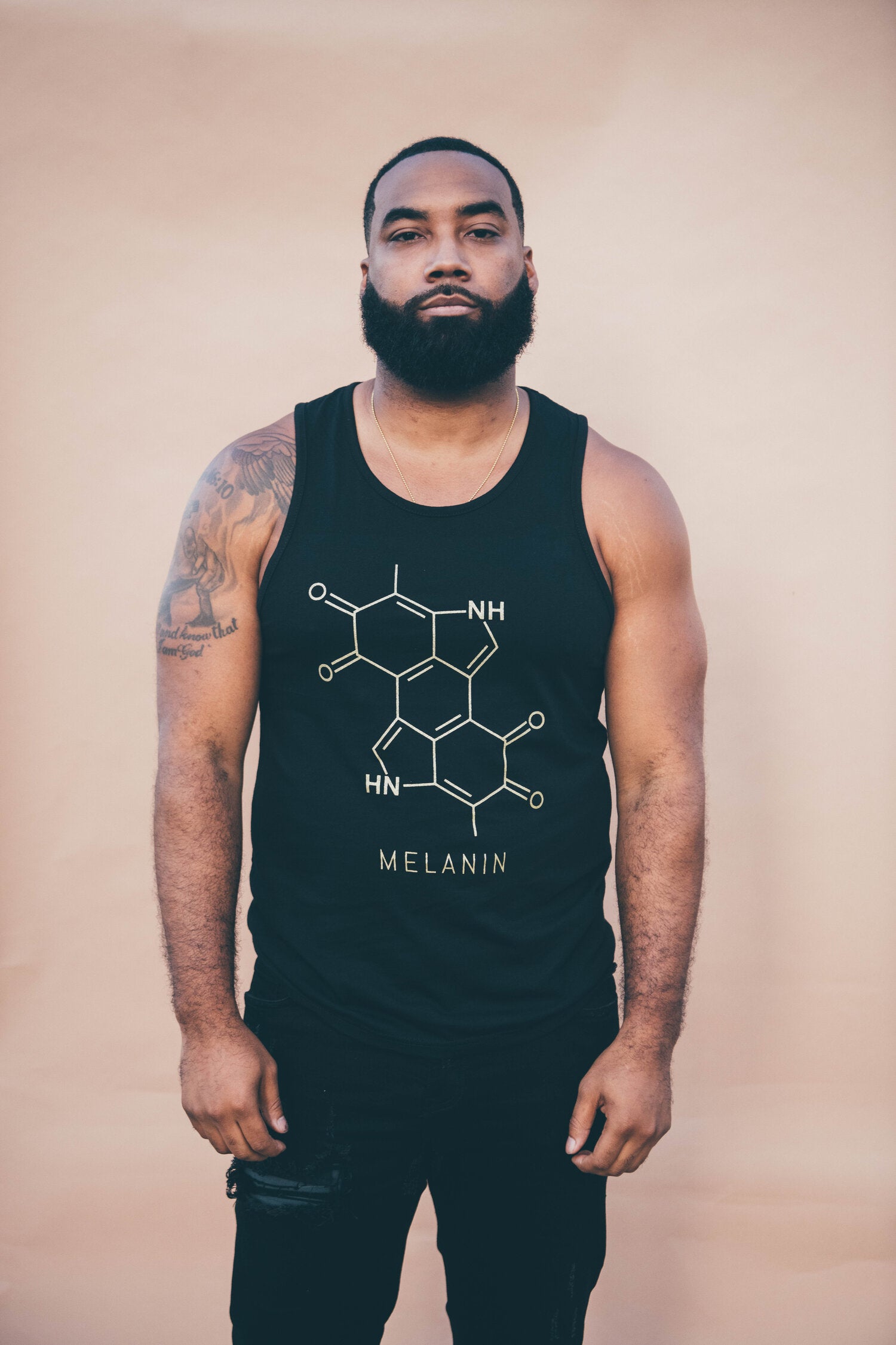 A man with a beard showcases the Men’s Melanin Tank, featuring a gold print of the melanin chemical structure. Clad in this stylish black top, his tattooed arms add character as he stands against a beige background.