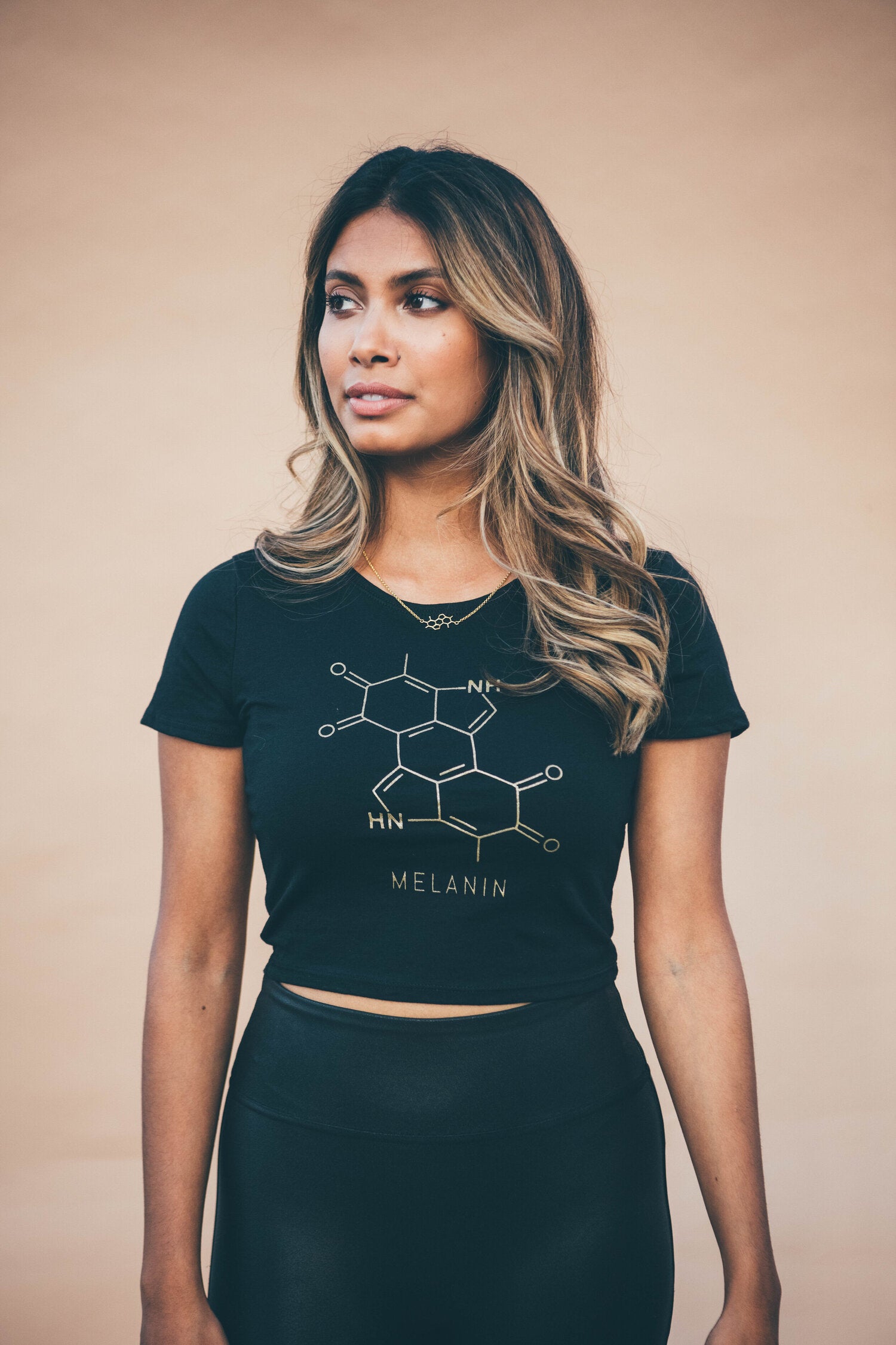 A person with long, wavy hair stands against a plain background wearing a womens cropped T-shirt that features a gold print of the chemical structure labeled Melanin. They look to the side with a neutral expression.