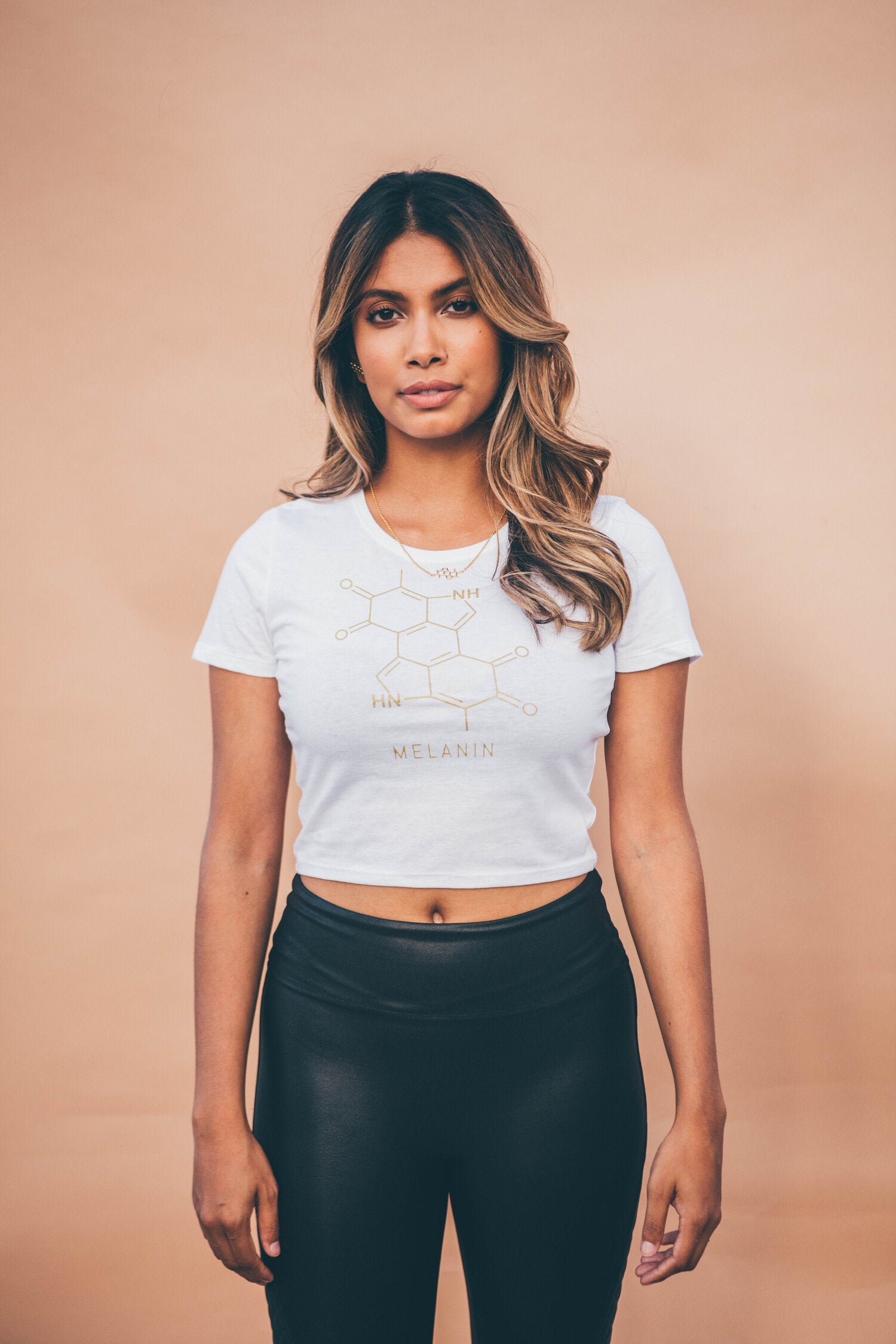 A woman stands against a neutral background wearing a white, women’s cropped t-shirt adorned with a gold print of the melanin molecule, paired perfectly with black leggings. Her hair cascades in loose waves.