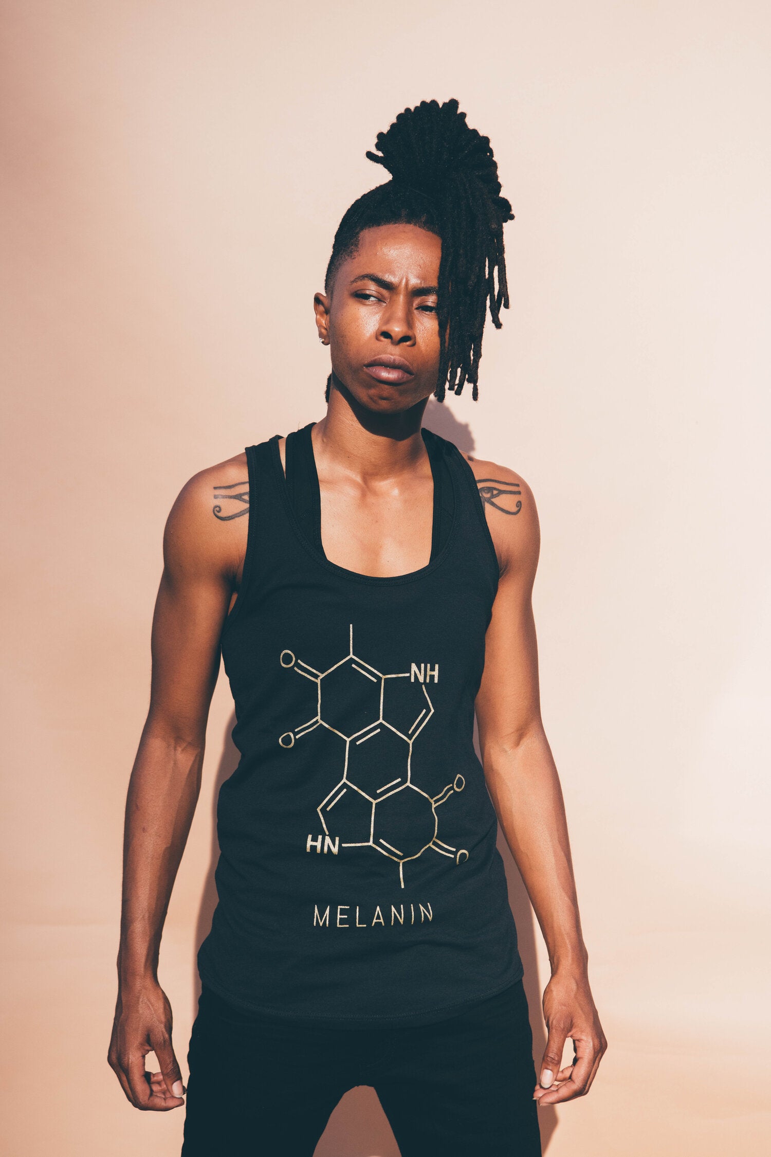 A woman in a black tank top with a striking gold print of the melanin molecule stands confidently against a beige background. She showcases long, styled hair and visible tattoos.