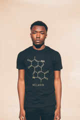 A person wearing a men’s black T-shirt adorned with a gold print of the chemical structure labeled Melanin stands against a neutral background. They have short hair and a neutral expression, adding to the shirts striking simplicity.