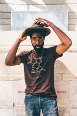 A man adjusts his wide-brimmed hat, showcasing a brown mens T-shirt adorned with a gold print of a chemical structure and the word Melanin. His beaded necklace gleams as sunlight dances across the engraved stone wall behind him.