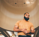 A man with a beard stands beneath a circular ceiling feature in a warmly lit building. Hes wearing a men’s T-shirt showcasing a melanin chemical structure, paired with black pants and a wristwatch, as he holds his hands together.