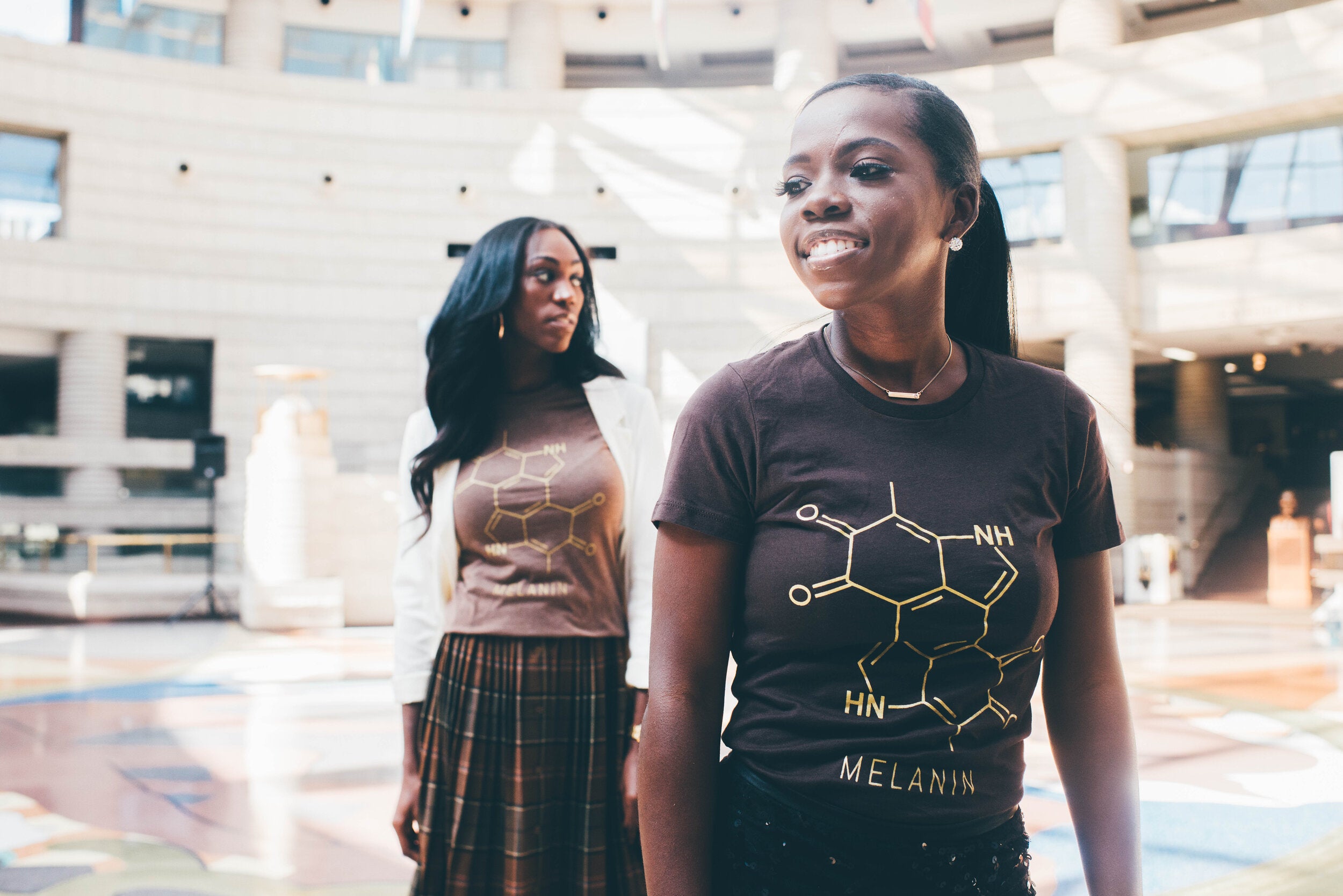 Two women wearing slim fit brown T-shirts featuring a melanin molecule design stand in a modern atrium. One looks forward smiling, while the other gazes to the side. Sunlight streams from above, illuminating the spacious, circular space.