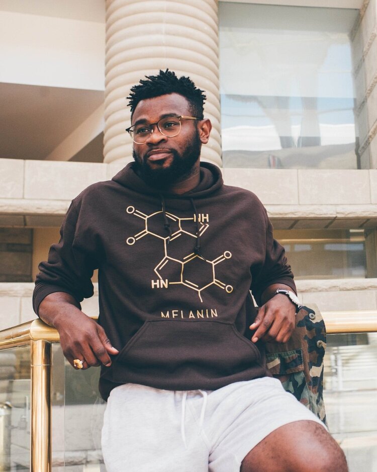 A person in glasses wears a brown unisex hoodie detailed with a molecular structure and the word MELANIN, paired with light gray shorts. They lean against a railing, set against the backdrop of a modern building.