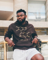 A person in glasses wears a brown unisex hoodie detailed with a molecular structure and the word MELANIN, paired with light gray shorts. They lean against a railing, set against the backdrop of a modern building.