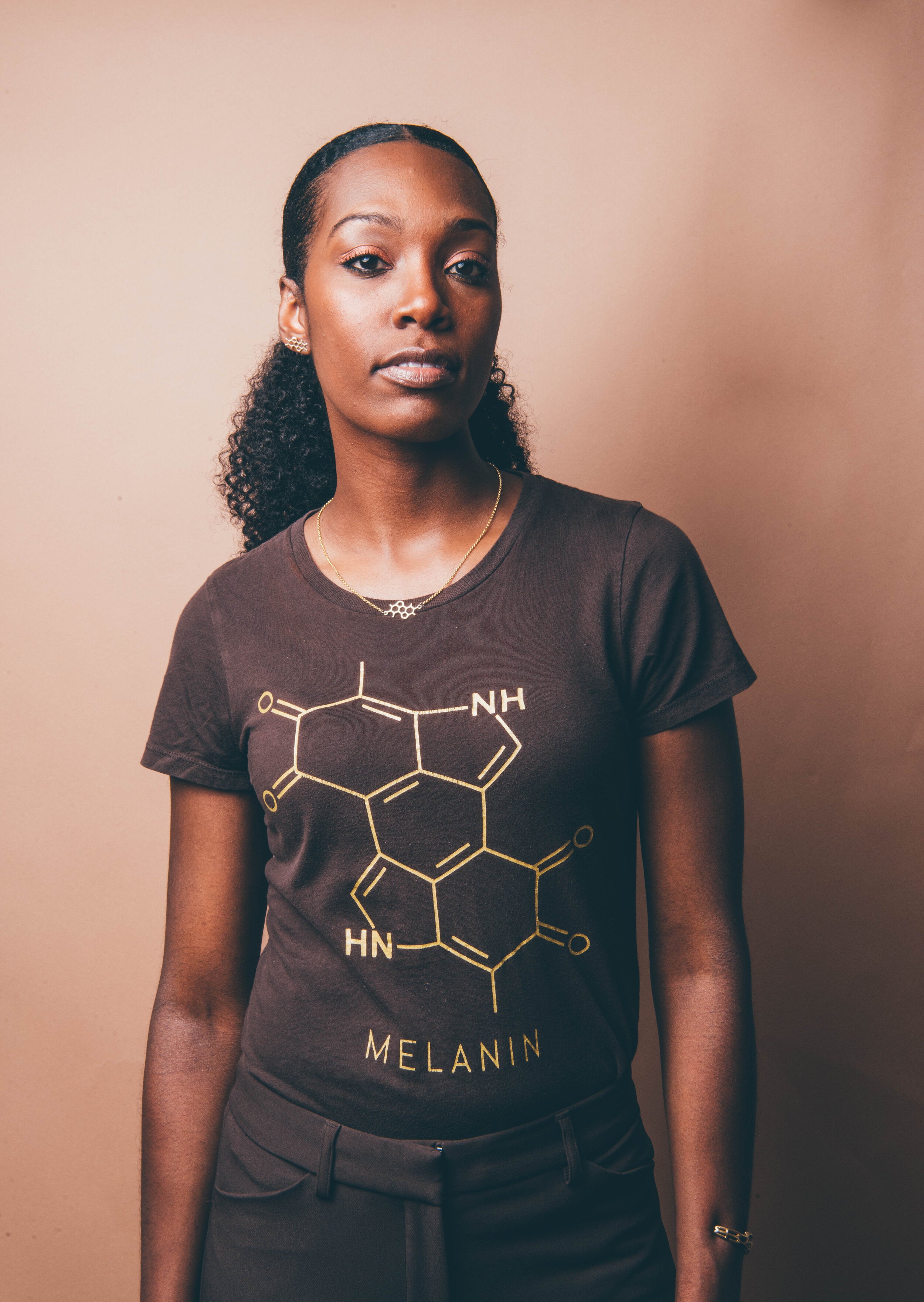 A person with curly hair stands confidently against a beige background, wearing a Womens T-shirt featuring a gold melanin chemical structure design, with the word MELANIN printed below.