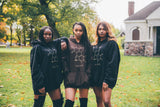 Three women strike a confident pose outdoors in unisex oversized hoodies featuring a striking melanin molecule design. They stand in front of a grassy area with trees and an elegant stone building, surrounded by leaves scattered on the ground.