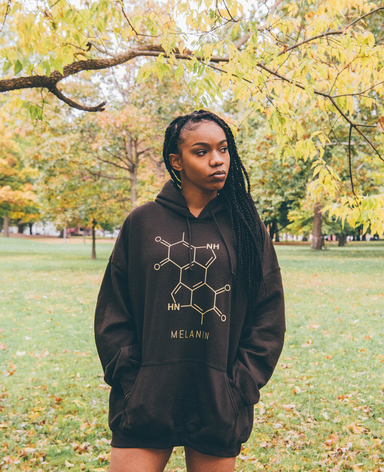 A person with long braided hair is wearing a black sweatshirt featuring a molecular design and the word MELANIN. Standing in a park with lush green grass and trees adorned with yellow leaves, their hands are casually tucked into the hoodie pockets.