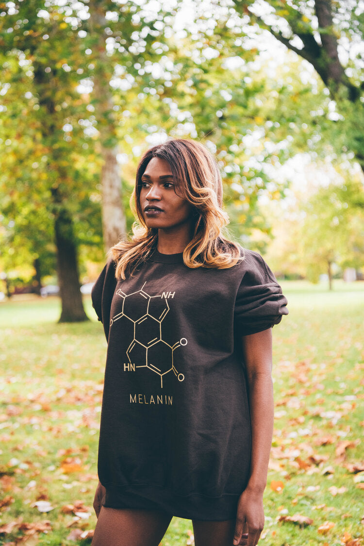 A person with long hair stands outdoors in a park, wearing a unisex crewneck sweatshirt adorned with the chemical structure of melanin. The background features green grass and trees with vibrant autumn leaves.