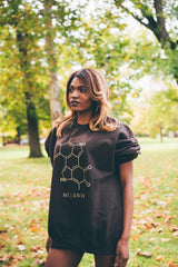 A person with long hair stands outdoors in a park, wearing a unisex crewneck sweatshirt adorned with the chemical structure of melanin. The background features green grass and trees with vibrant autumn leaves.
