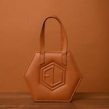 The HEX Mini Bag, a testament to quality craftsmanship, is hexagon-shaped and crafted from rich brown leather. It boasts two sturdy handles and a raised geometric design, perfectly complemented by the wooden surface and matching brown backdrop.