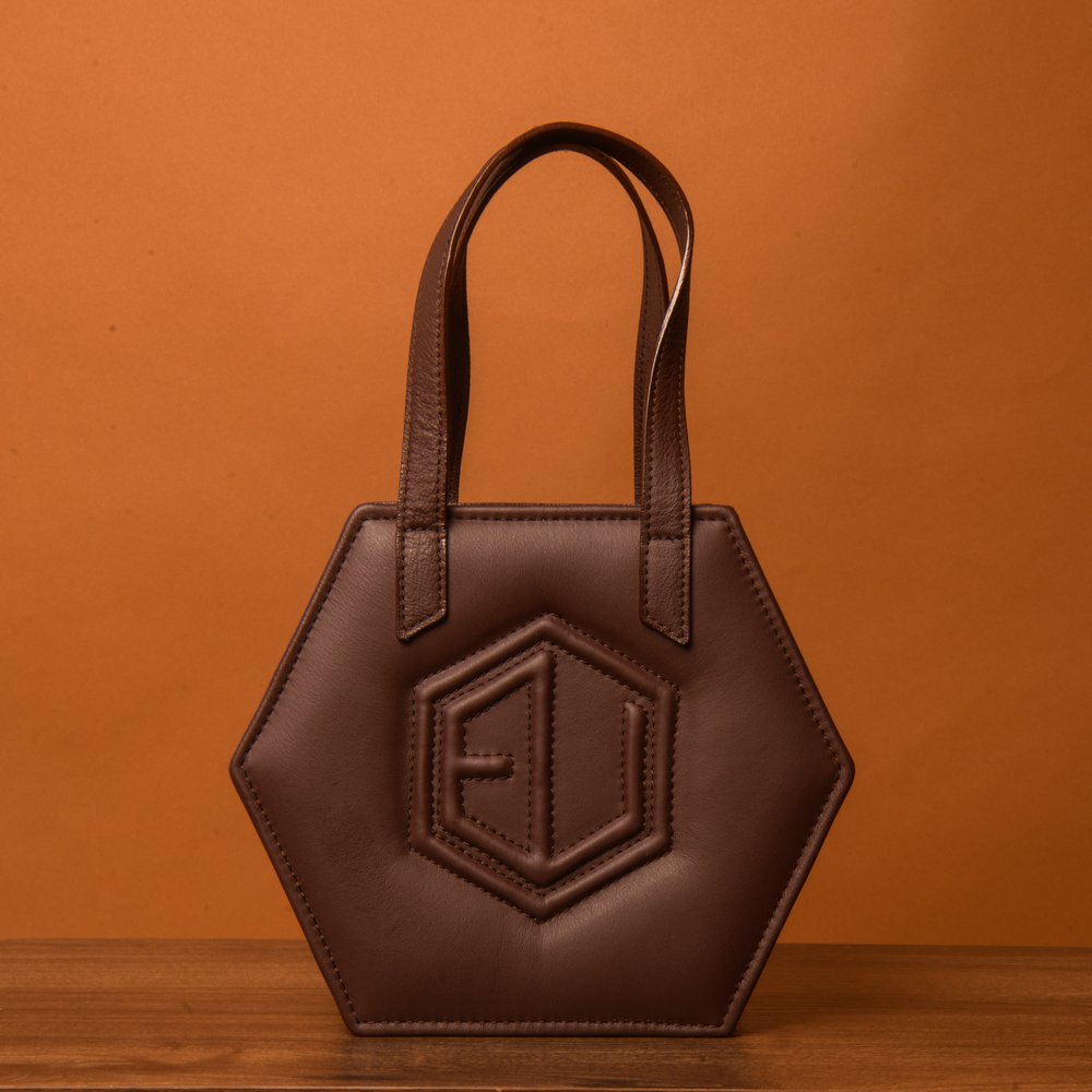 A HEX Mini Bag crafted in hexagonal brown leather, showcasing two sturdy handles and a raised geometric design at its core. Its elegantly displayed against a warm, orange-brown backdrop on a polished wooden surface.