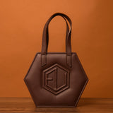 A HEX Mini Bag crafted in hexagonal brown leather, showcasing two sturdy handles and a raised geometric design at its core. Its elegantly displayed against a warm, orange-brown backdrop on a polished wooden surface.