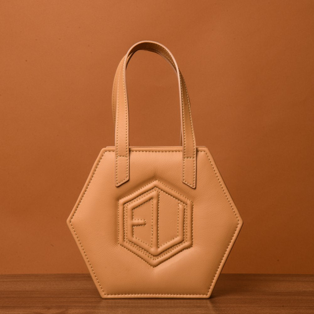 A beige hexagonal handbag, known as the HEX Mini Bag, with short handles is set against a matching brownish backdrop. It showcases a raised geometric pattern on the front, resembling eumelanin’s intricate design.