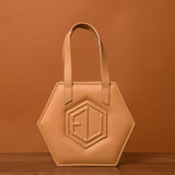 A beige hexagonal handbag, known as the HEX Mini Bag, with short handles is set against a matching brownish backdrop. It showcases a raised geometric pattern on the front, resembling eumelanin’s intricate design.