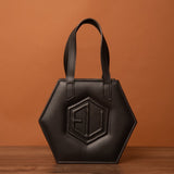 The HEX Mini Bag, a hexagonal black handbag with dual handles, showcases a raised geometric design on the front. It rests elegantly on a wooden surface, set against a warm brown background.