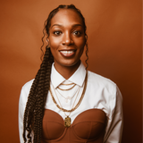 A person smiling against a brown background showcases their eumelanin-rich skin. Their long braided hair complements a white shirt and brown top, while multiple gold chains gracefully adorn their neck, creating a chic look that would be perfect for any trendy shop setting.