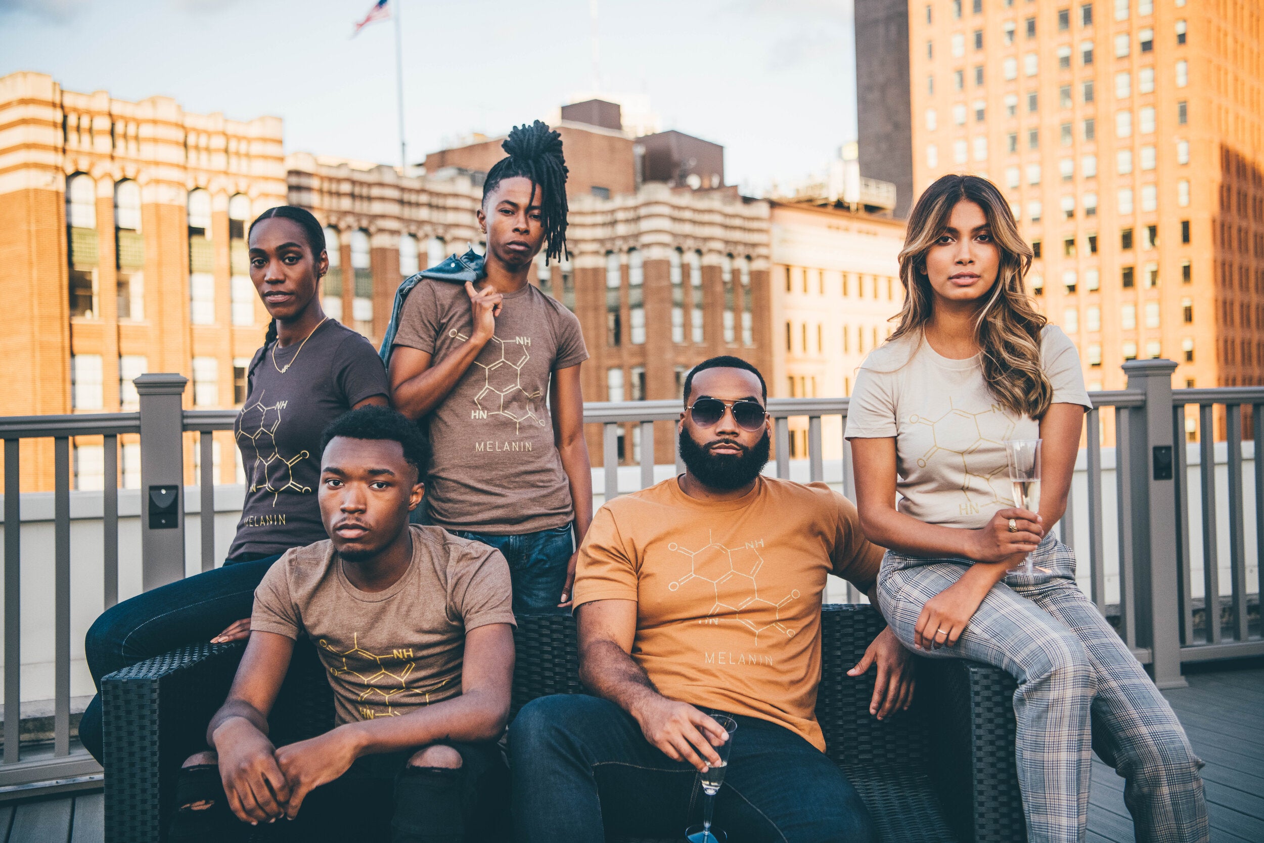 Five people sit and stand on a rooftop, city buildings framing their pose in the warm evening light. Clad in matching womens T-shirts adorned with melanins chemical structure, they exude confidence while shadows dance around them.