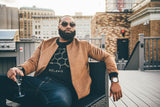 A man with a beard and sunglasses lounges on a rooftop deck. Sporting a tan jacket over his black sweatshirt adorned with the chemical structure of melanin, he holds a glass, cityscape stretching behind him and outdoor barbecue equipment close by.
