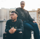 Two people are casually sitting on a rooftop with city buildings in the background. The person on a chair wears a black crop hoodie, while the one seated on the ground sports a black sweater with a molecular design. They appear relaxed under the evening sky.