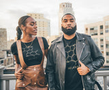 A man and woman stand on an urban rooftop, showcasing their unique style. Sporting T-shirts with a eumelanin molecule design, she pairs hers with a brown leather skirt and necklace, while he opts for a leather jacket. City buildings line the backdrop under the cloudy sky—an ideal scene to shop this chic look.