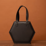 A sleek hexagonal black HEX Bag with two sturdy handles rests elegantly on a wooden surface. The brand name is embossed prominently on the front, standing out against the bags design. It sits against a warm, earthy orange background, enhancing its stylish allure.