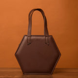 The HEX Mini Bag, a hexagon-shaped brown leather handbag with dual handles, stands out against a warm, orange-brown background.
