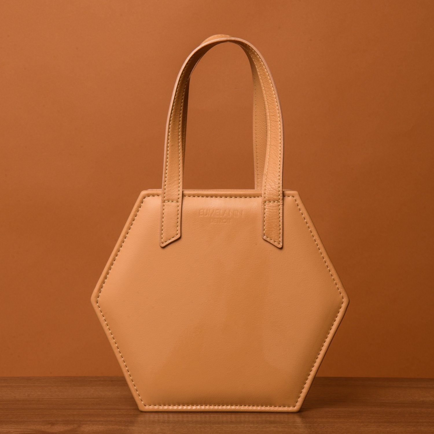 A beige, hexagonal HEX Mini Bag with two straps stands on a wooden surface against a brown background.