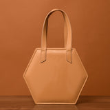A beige, hexagonal HEX Mini Bag with two straps stands on a wooden surface against a brown background.