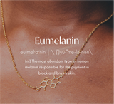Close-up of a person wearing a gold necklace featuring a melanin molecule pendant. The text Eumelanin appears with its phonetic spelling and definition, highlighting it as the pigment behind black and brown skin tones—a unique piece you can shop to celebrate science in style.