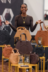 A person stands smiling behind a display of hexagonal handbags in various colors, including black, brown, and tan at the Eumelanin Shop. The bags are arranged on yellow stands and paired with photographs of models. The person wears a dark outfit and gold jewelry.