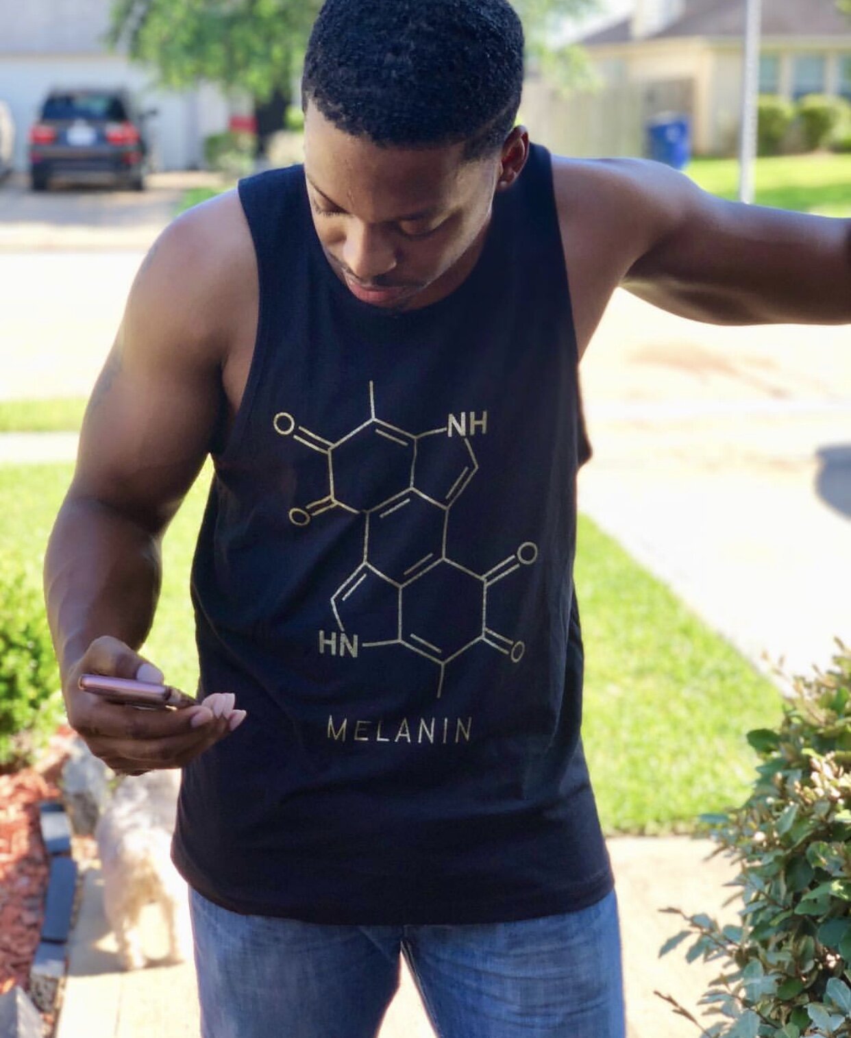 A man in a Mens Melanin Tank, featuring a melanin structure in gold print, looks down at his phone. He stands outdoors under the sunlight, with a green lawn and houses serving as the background.