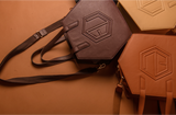 Four hexagonal leather handbags in earthy brown and tan shades lay on a brown surface. Each features embossed geometric patterns and adjustable straps, elegantly arranged in a semi-circle, inviting you to shop the unique eumelanin-inspired designs.