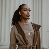 A woman stands confidently in a brown coat and a scientific-themed t-shirt with molecule illustrations. Her curly eumelanin-rich hair is tied back, complementing her gold hoop earrings and pendant necklace. Behind her, large white pillars add to the ambiance of this sophisticated shop.