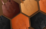 Geometric HEX Bags in shades of brown, orange, and black with embossed logos are arranged on a brown background. Featuring premium hardware and luxe allure leather, the bags hexagonal shapes contrast stylishly, showcasing a modern design.
