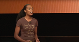 A woman with long, curly hair is speaking on stage. She wears a brown T-shirt featuring the chemical structure labeled Eumelanin. The background is a warm, wood-paneled wall. Her gestures are animated, as if inviting the audience to shop for knowledge and insight.