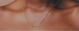 A person wearing a gold necklace from Eumelanin Shop, featuring an abstract, honeycomb-shaped pendant. The necklace rests elegantly on their collarbone against a neutral background.