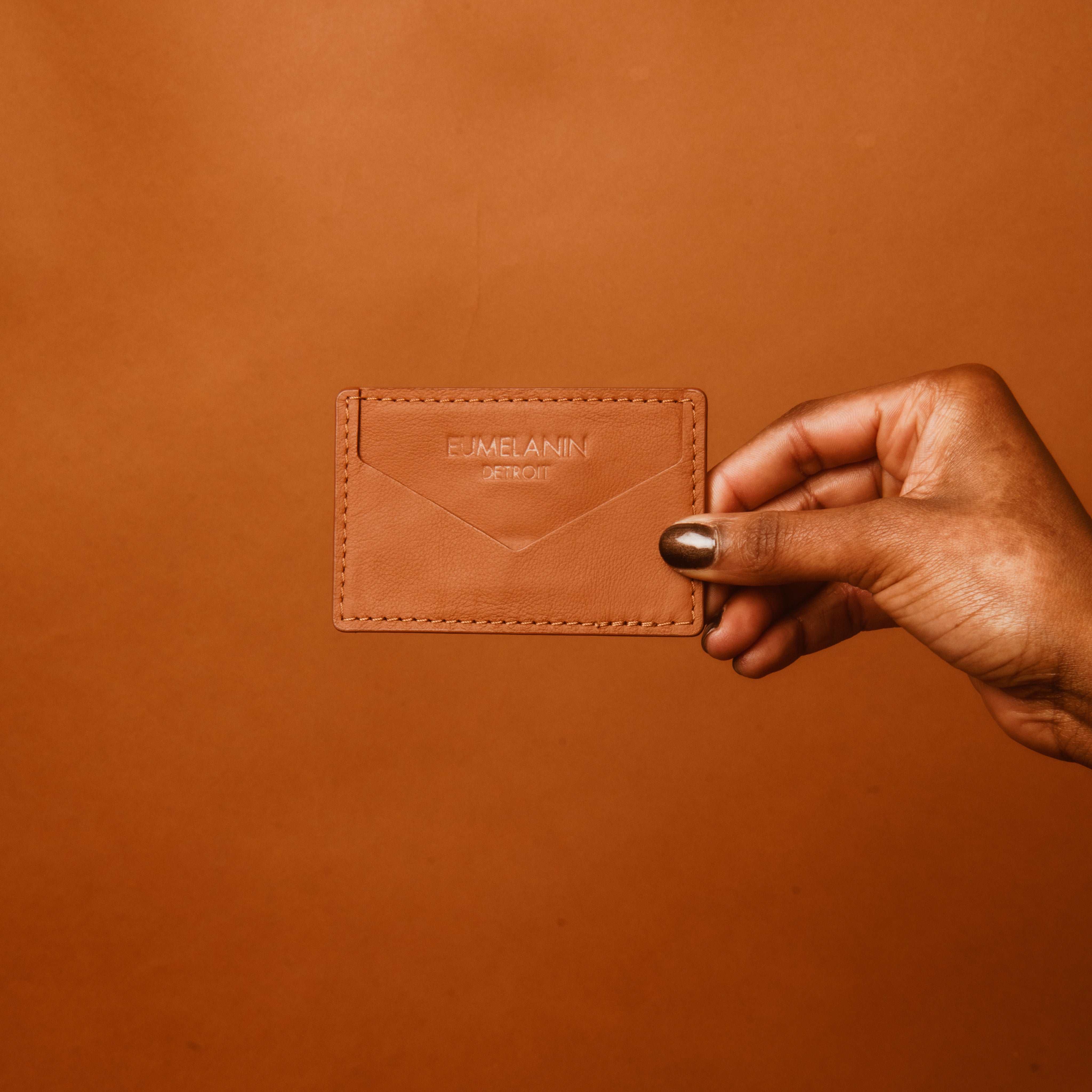 A hand holding a tan leather cardholder from EUMELANIN DETROIT, ideal for the fashion-savvy shopper, is showcased against a matching tan background.