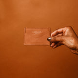 A hand holding a tan leather cardholder from EUMELANIN DETROIT, ideal for the fashion-savvy shopper, is showcased against a matching tan background.