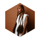 A woman with long braided hair, rich in eumelanin, stands in profile against a brown background. She wears a sleeveless white jacket and layered necklaces, captured within a hexagonal frame.