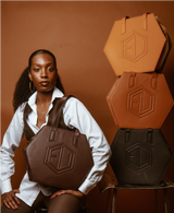 A person with styled eumelanin-rich hair poses with a brown hexagonal handbag. Next to them is a stack of three more bags in orange, tan, and black. They stand against a brown background, wearing a white shirt and dark pants, showcasing the perfect shop-ready ensemble.