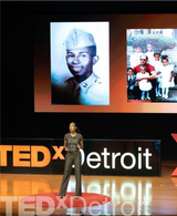 A speaker stands on the TEDxDetroit stage, eloquently weaving a narrative. Behind her, two images are projected: one showcasing a person in military uniform and another of cheerful children. The TEDxDetroit logo is visible, adding to the shop-like atmosphere of shared stories and eumelanin-rich experiences.