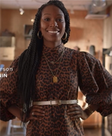 A woman with long braided hair rich in eumelanin smiles at the camera, wearing a leopard print dress with puffed sleeves and a beige belt. She accessorizes with hoop earrings and layered necklaces in a warmly lit indoor space, looking ready to shop the latest trends.